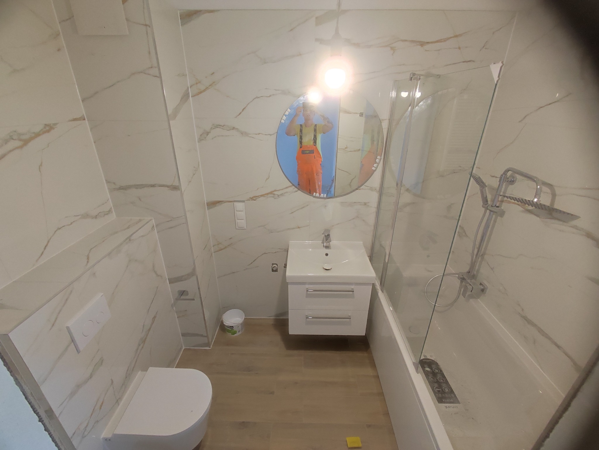 Nowoczesna łazienka z marmurowymi płytkami, kabiną prysznicową za szkłem, białą umywalką z szafką i wiszącą toaletą; w lustrze odbija się instalator w pomarańczowych spodniach.