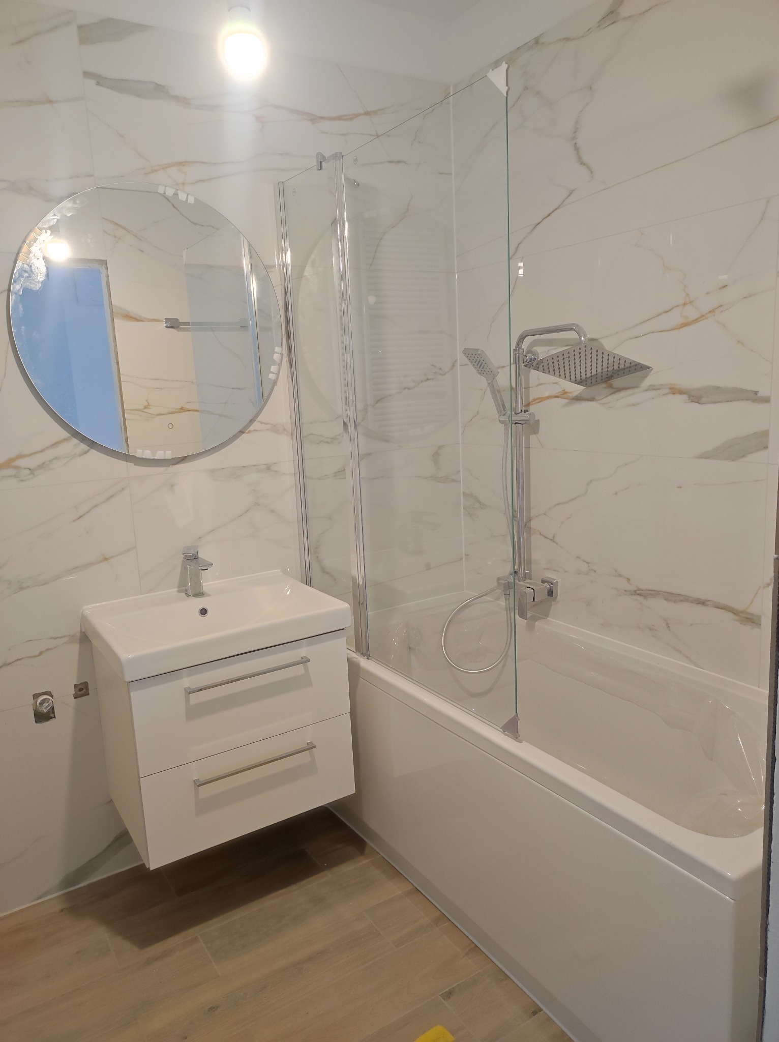 Nowoczesna łazienka z marmurowymi płytkami, okrągłym lustrem z podświetleniem LED, białą szafką pod umywalką z dwoma szufladami i kabiną prysznicową typu walk-in z deszczownicą.