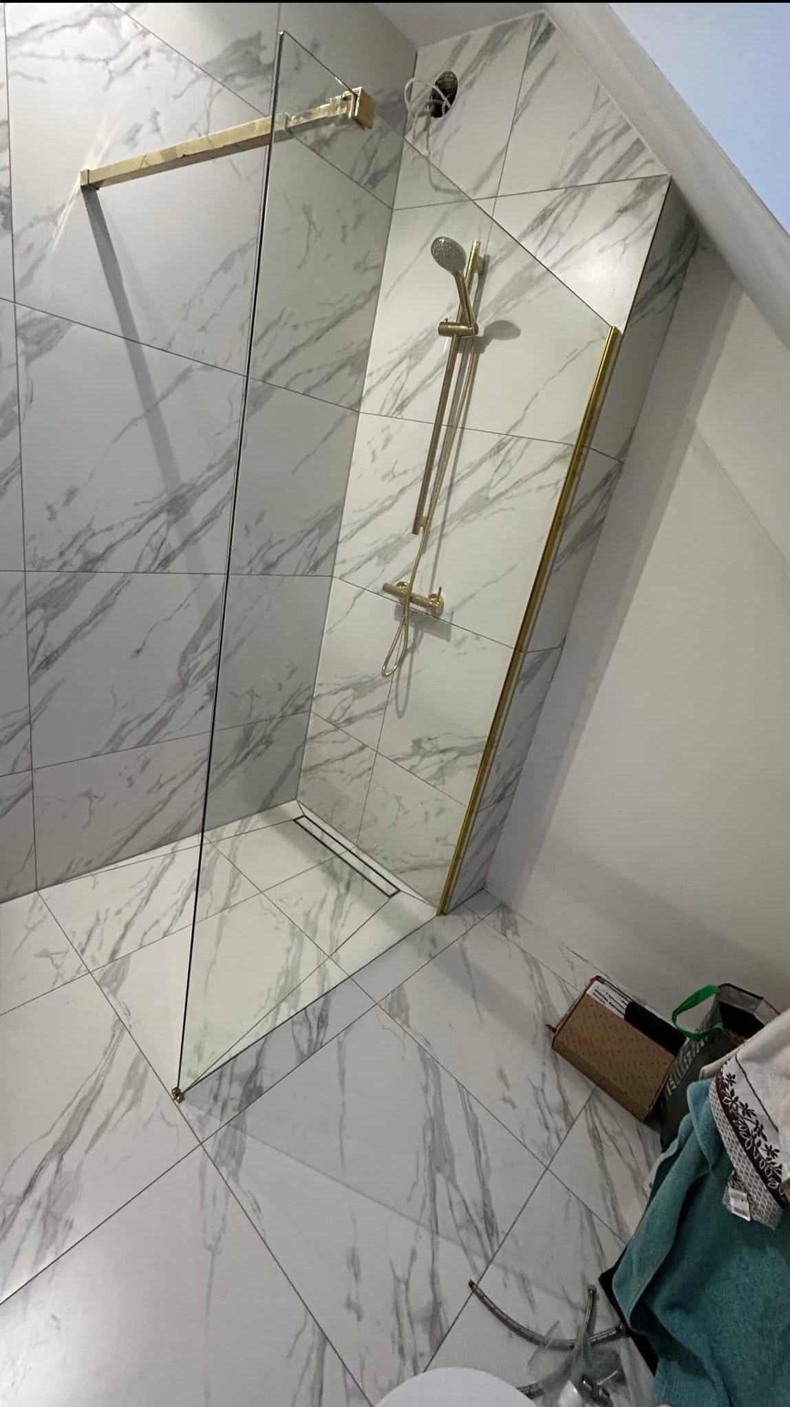 Kabina prysznicowa typu walk-in z marmurowymi płytkami, złotymi wykończeniami i odpływem liniowym, częściowo widoczne akcesoria łazienkowe w rogu.