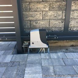 D.O. SERWIS DEMIAN OLAR - Szary automat do bramy przesuwnej zamontowany na szynie, widok z bliska na kostce brukowej przy szarym ogrodzeniu i ścianie z szarej cegły.