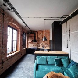 D.O. SERWIS DEMIAN OLAR - Loftowe wnętrze z odsłoniętą cegłą i instalacją elektryczną natynkową w czarnych rurkach, widok na kuchnię i turkusową sofę.