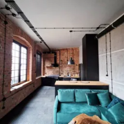 Loftowe wnętrze z odsłoniętą cegłą i instalacją elektryczną natynkową w czarnych rurkach, widok na kuchnię i turkusową sofę.
