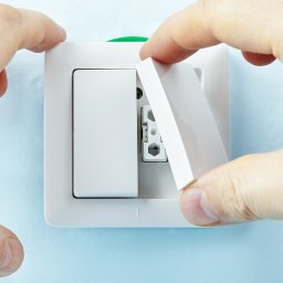 D.O. SERWIS DEMIAN OLAR - Instalacja białego włącznika światła na błękitnej ścianie, widoczne dłonie montera zakładające osłonę.