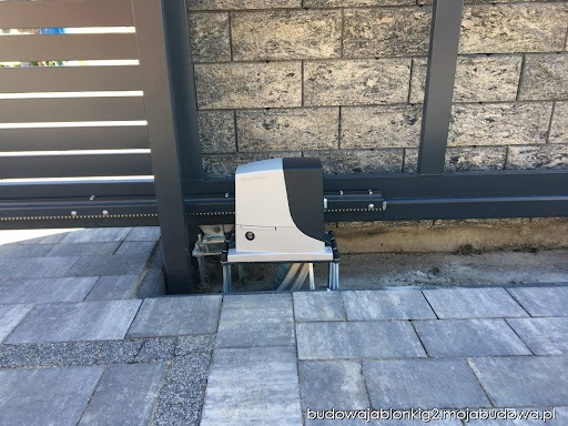 Szary automat do bramy przesuwnej zamontowany na szynie, widok z bliska na kostce brukowej przy szarym ogrodzeniu i ścianie z szarej cegły.