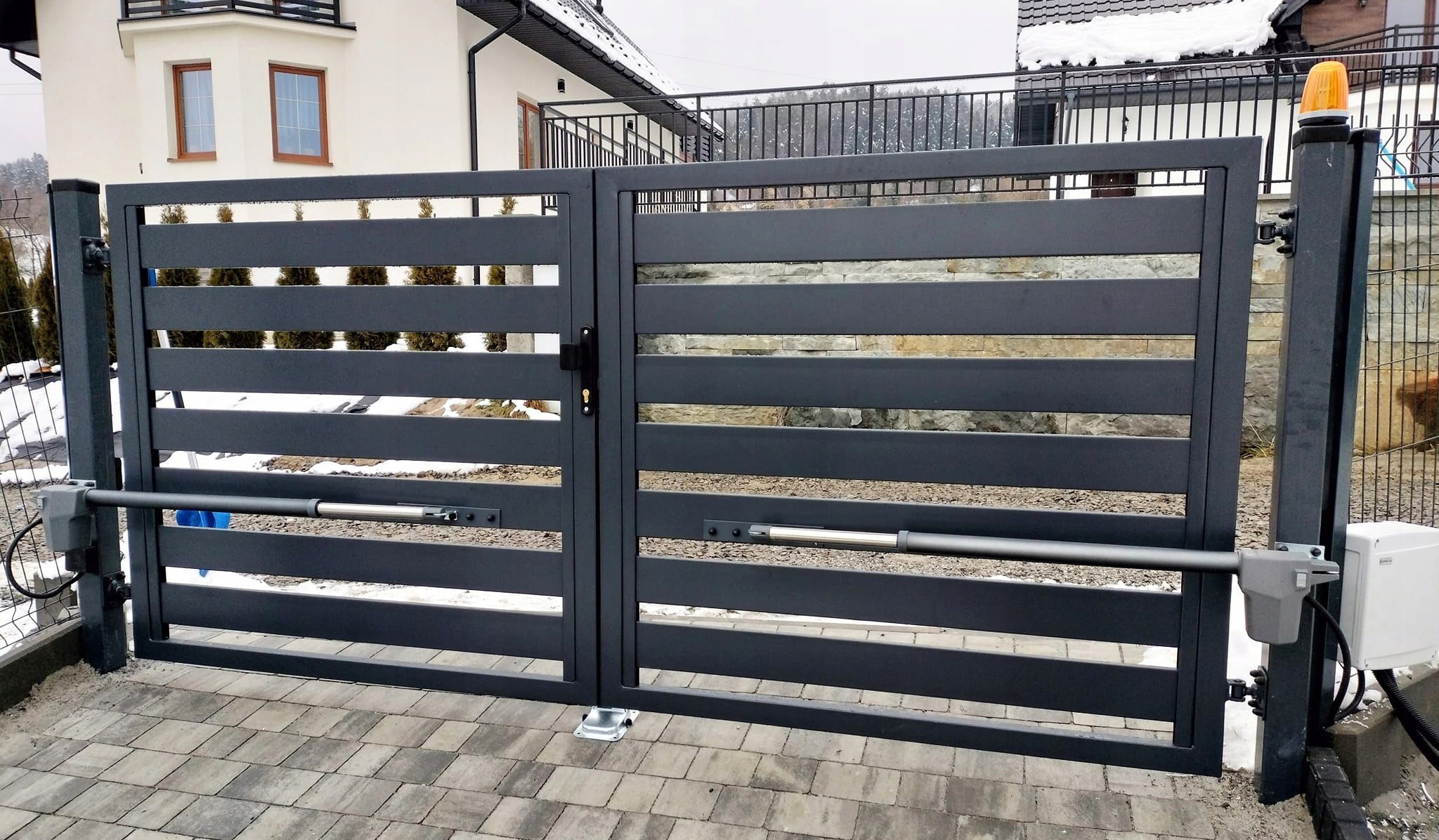 Nowoczesna, grafitowa brama dwuskrzydłowa z automatyką zamontowaną na słupkach, widoczny napęd liniowy i skrzynka sterująca, kostka brukowa na podjeździe, dom jednorodzinny w tle.