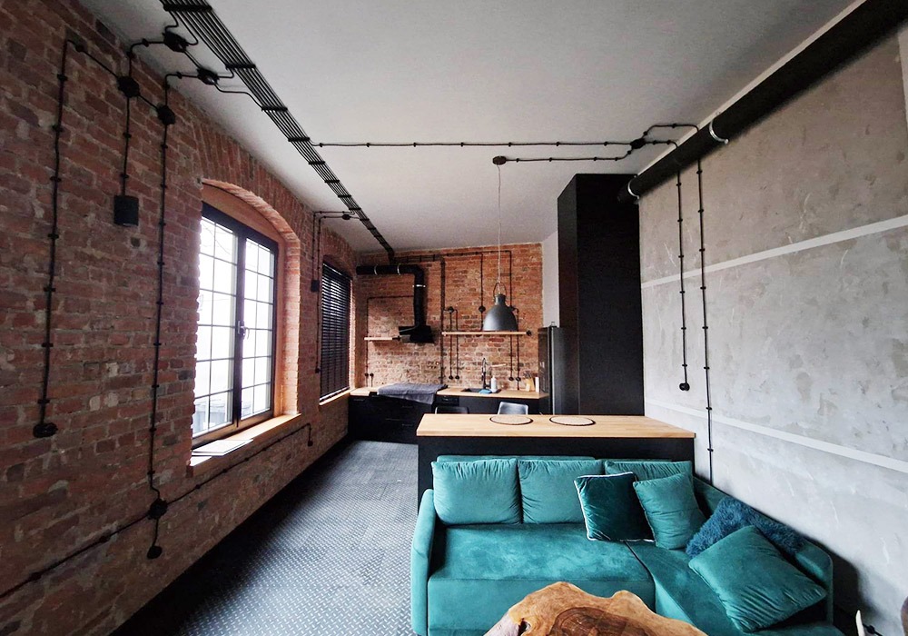 Loftowe wnętrze z odsłoniętą cegłą i instalacją elektryczną natynkową w czarnych rurkach, widok na kuchnię i turkusową sofę.