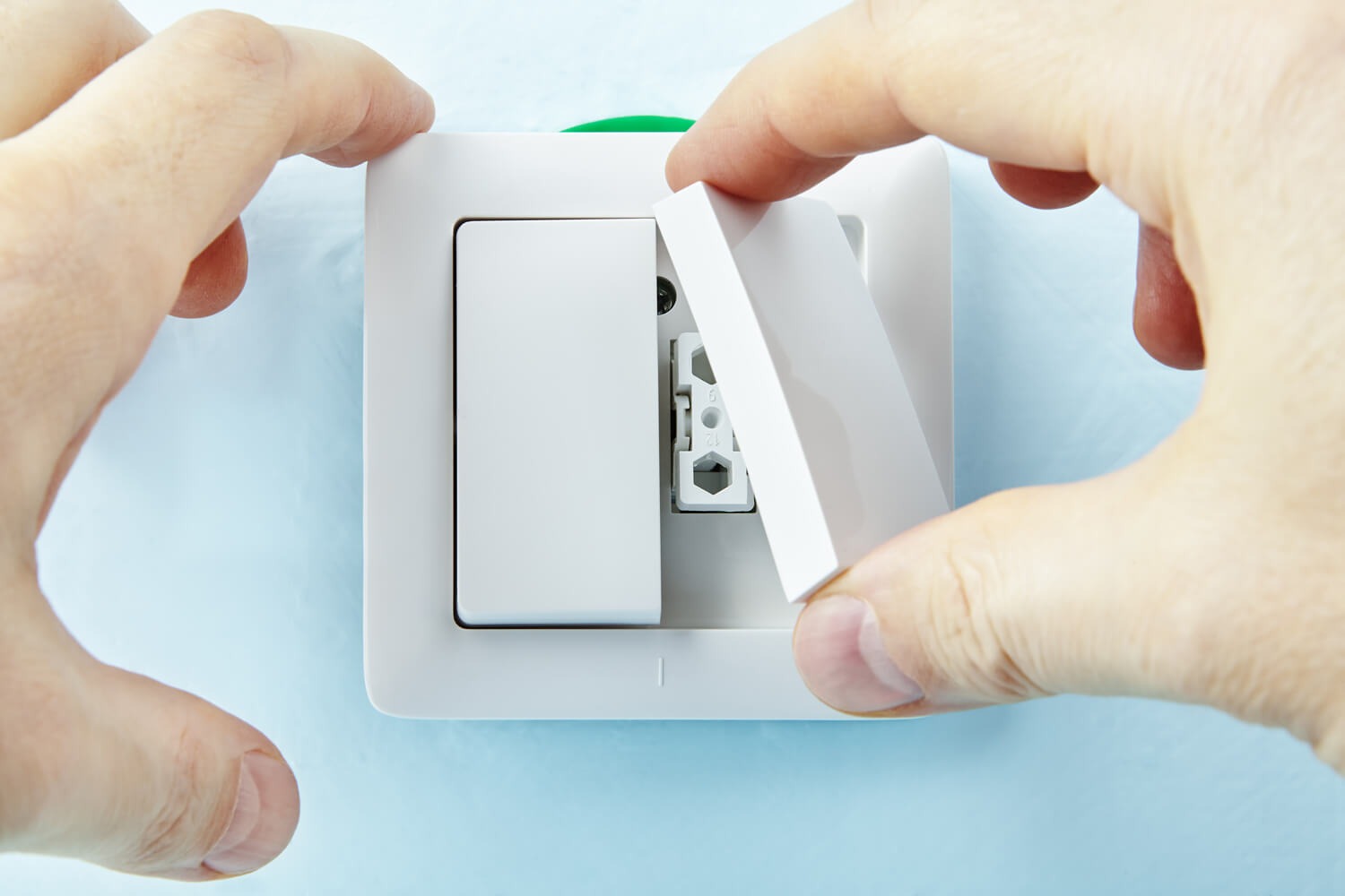 Instalacja białego włącznika światła na błękitnej ścianie, widoczne dłonie montera zakładające osłonę.