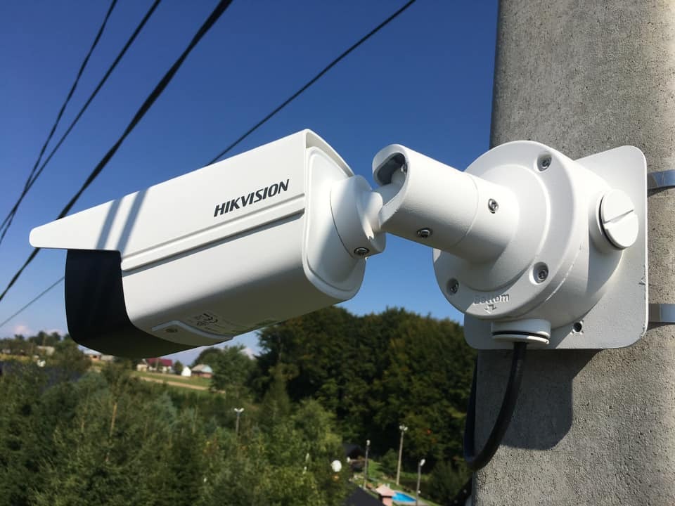 Zewnętrzna kamera monitoringu Hikvision zamontowana na betonowym słupie, widok z dołu na tle błękitnego nieba i zieleni drzew.