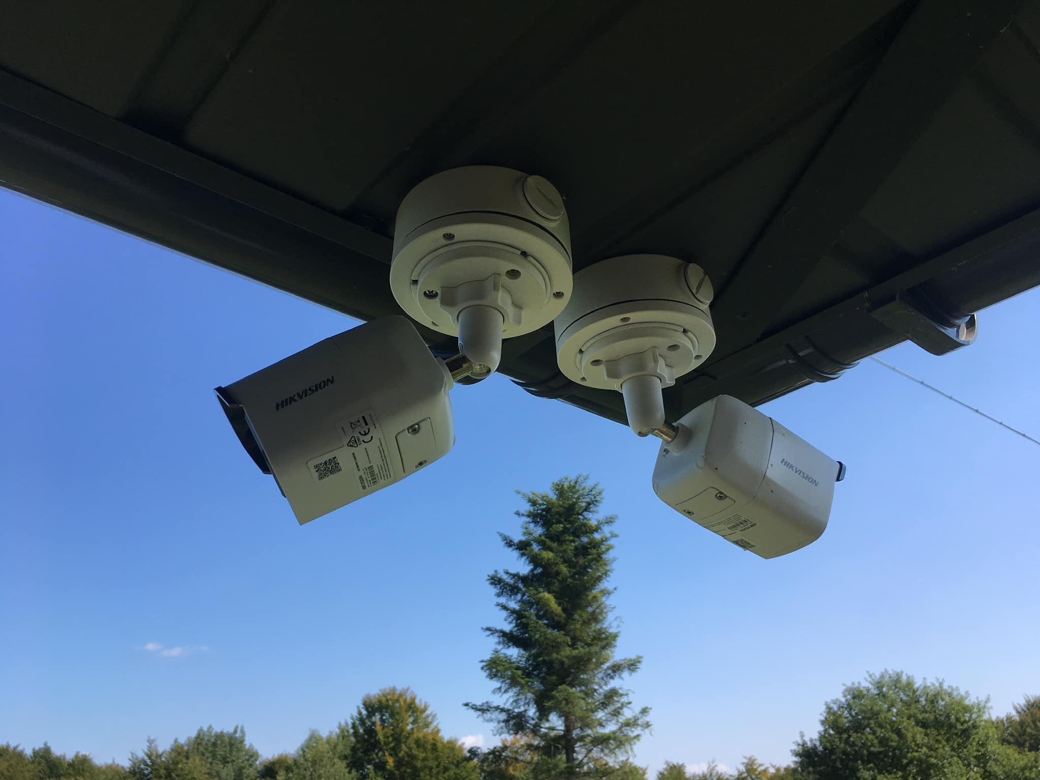 Dwie kamery monitoringu Hikvision zamontowane pod zadaszeniem, widok na błękitne niebo i drzewa.