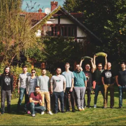 Grupa kilkunastu osób, prawdopodobnie zespół, pozuje na trawniku przed domem z czerwonym dachem, dwie osoby trzymają nad głowami duże cukinie.