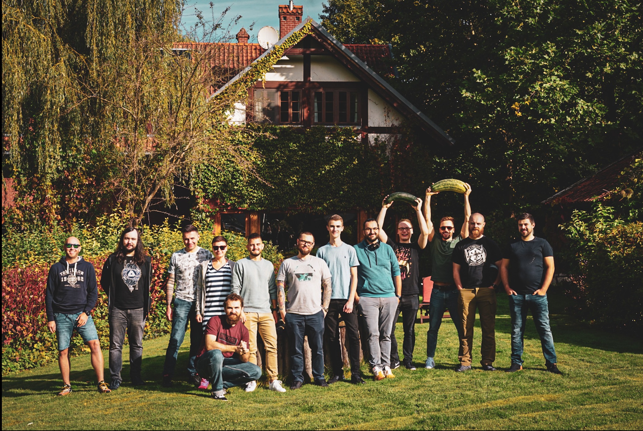 Grupa kilkunastu osób, prawdopodobnie zespół, pozuje na trawniku przed domem z czerwonym dachem, dwie osoby trzymają nad głowami duże cukinie.