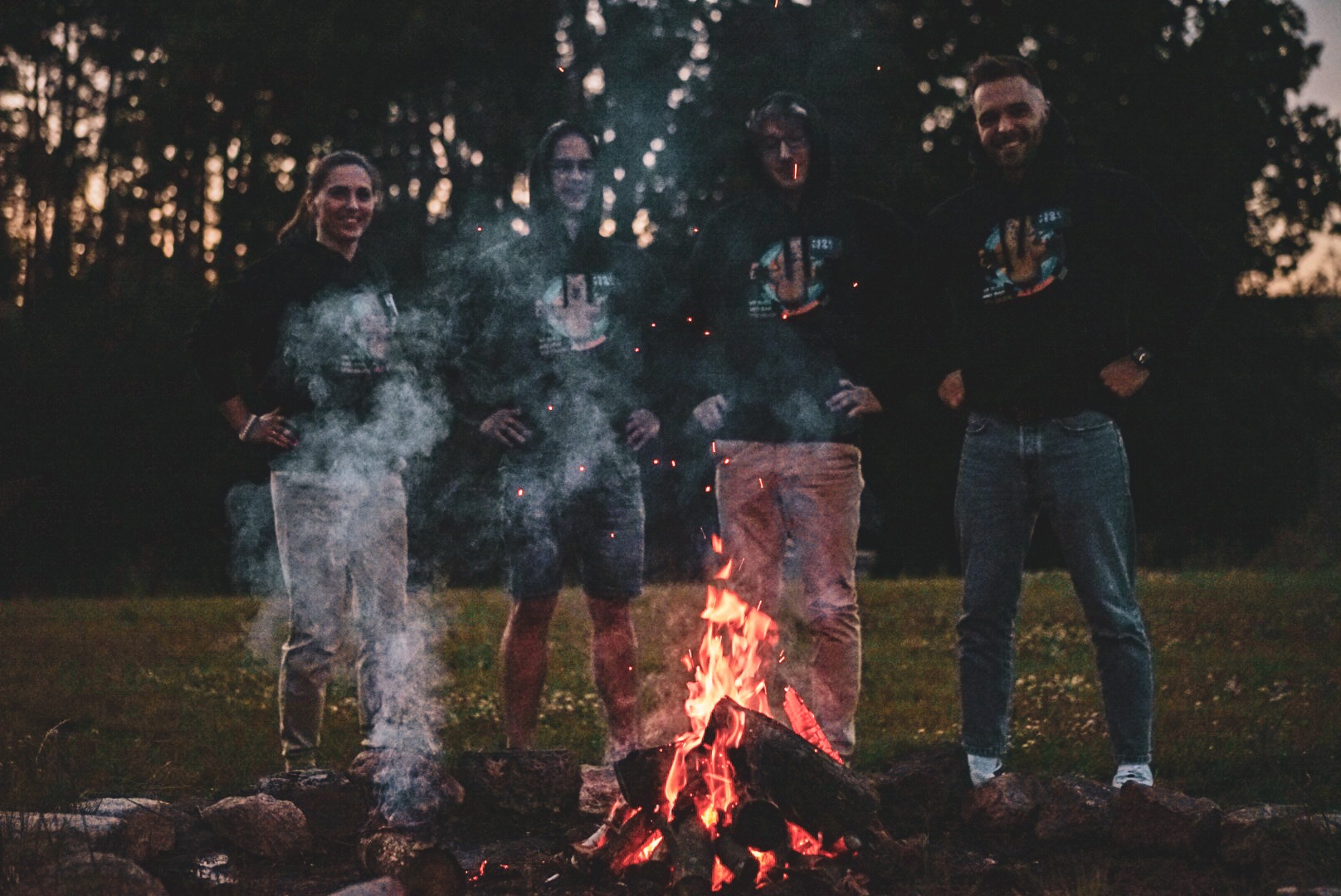 Czteroosobowy zespół w ciemnych bluzach z logo firmy, stojący wokół ogniska w plenerze, z dymem unoszącym się w powietrzu, sugerujący integrację zespołu IT.