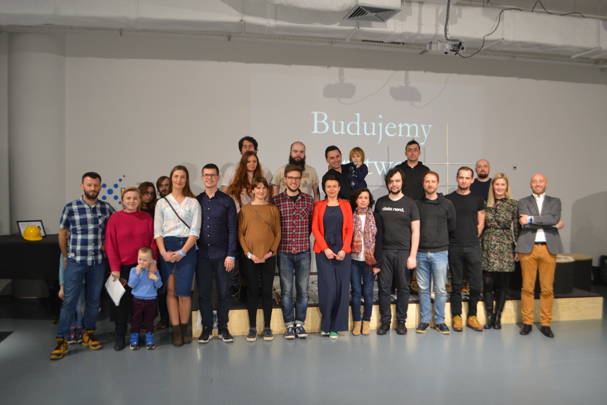 Grupa około dwudziestu osób, w tym dziecko, stoi na tle ściany z wyświetlanym napisem 'Budujemy' oraz dwoma projektorami. Niektóre osoby ubrane są w koszulki z napisami związanymi z danymi.