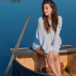 Młoda kobieta w białej bluzce siedzi w łodzi na spokojnym jeziorze, z wiosłem opartym o burtę, delikatne światło podkreśla jej naturalną urodę.