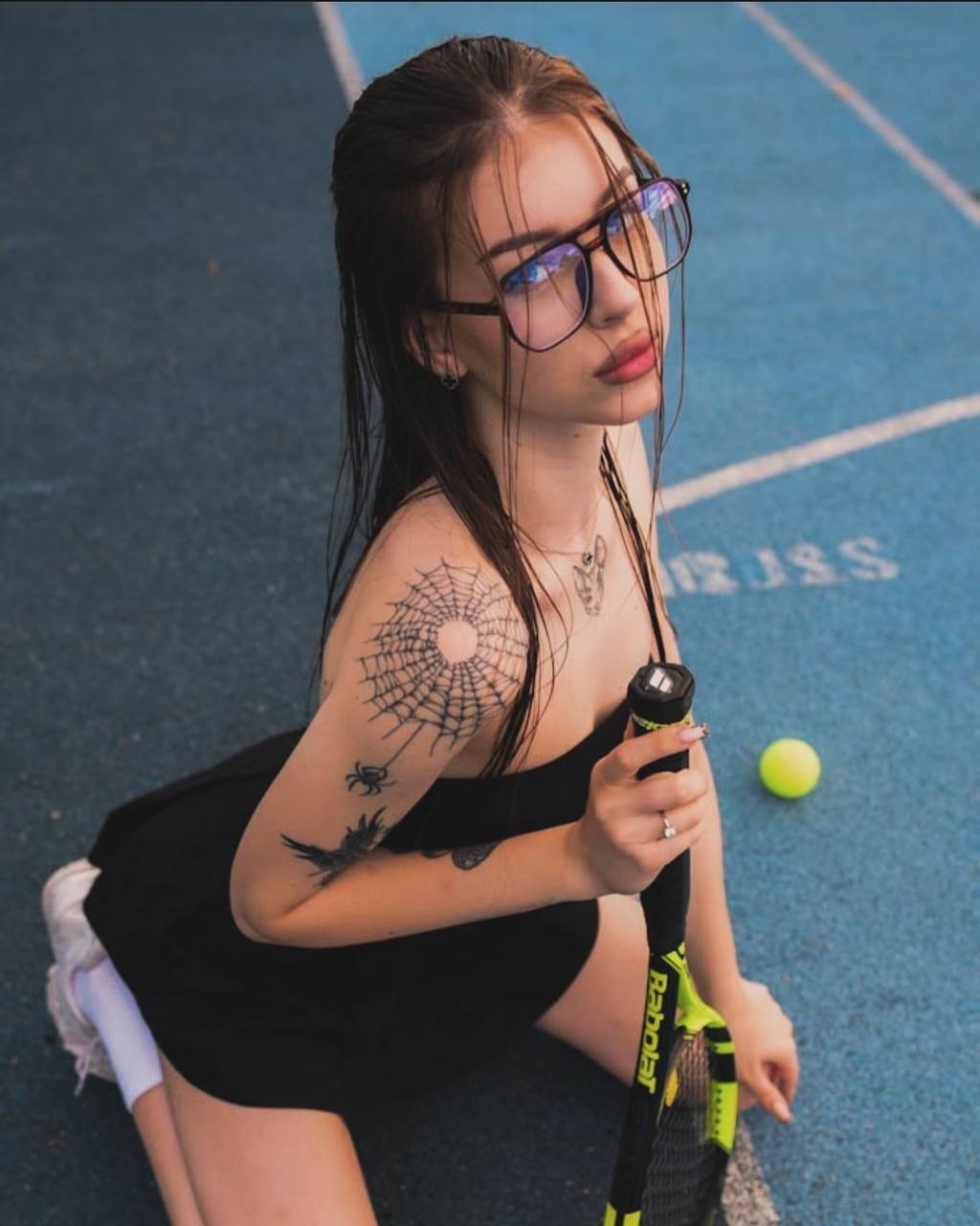 Młoda kobieta w okularach, z tatuażami na ramieniu, siedzi na niebieskim korcie tenisowym, trzyma rakietę Babolat i patrzy w górę.
