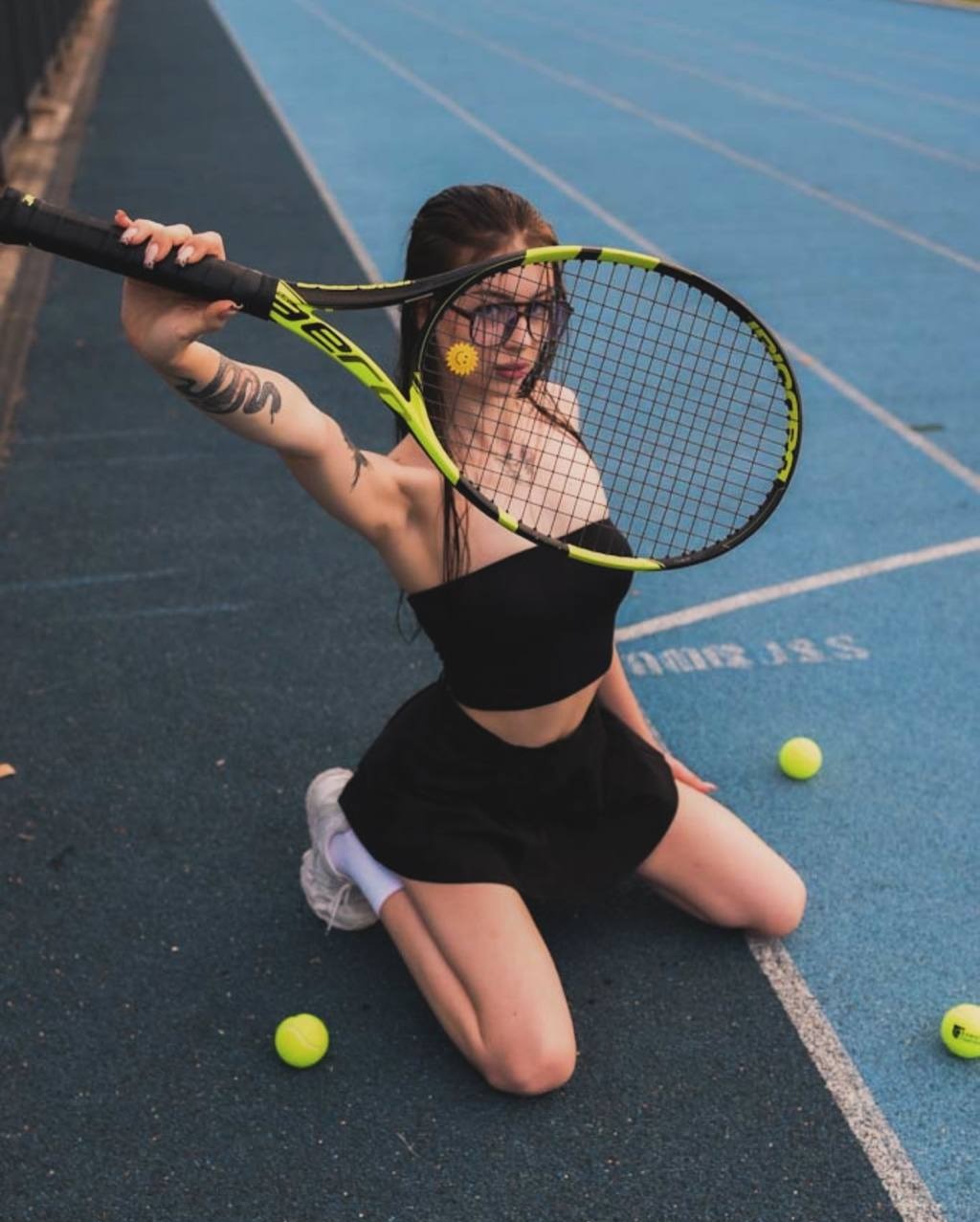 Kobieta w okularach klęczy na niebieskim bieżni z rakietą tenisową i piłkami, z tatuażem węża na ramieniu i naklejką słonecznika na twarzy.