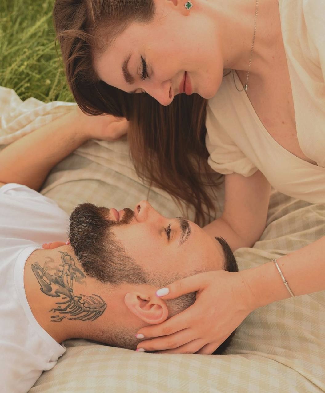 Para leżąca na kocu w trawie, kobieta opiera się nad mężczyzną, delikatnie trzymając jego głowę w dłoniach. Mężczyzna ma tatuaż na szyi.