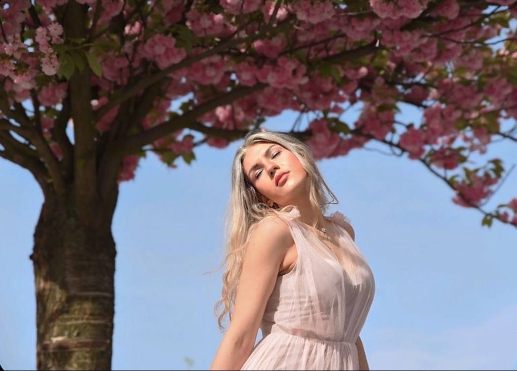 Młoda kobieta w zwiewnej, jasnoróżowej sukience pozuje pod kwitnącym drzewem wiśni o różowych kwiatach na tle błękitnego nieba.