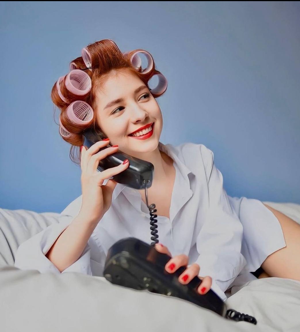 Uśmiechnięta kobieta z lokówkami na głowie, w białej koszuli, leżąca na łóżku i rozmawiająca przez czarny, stary telefon stacjonarny.