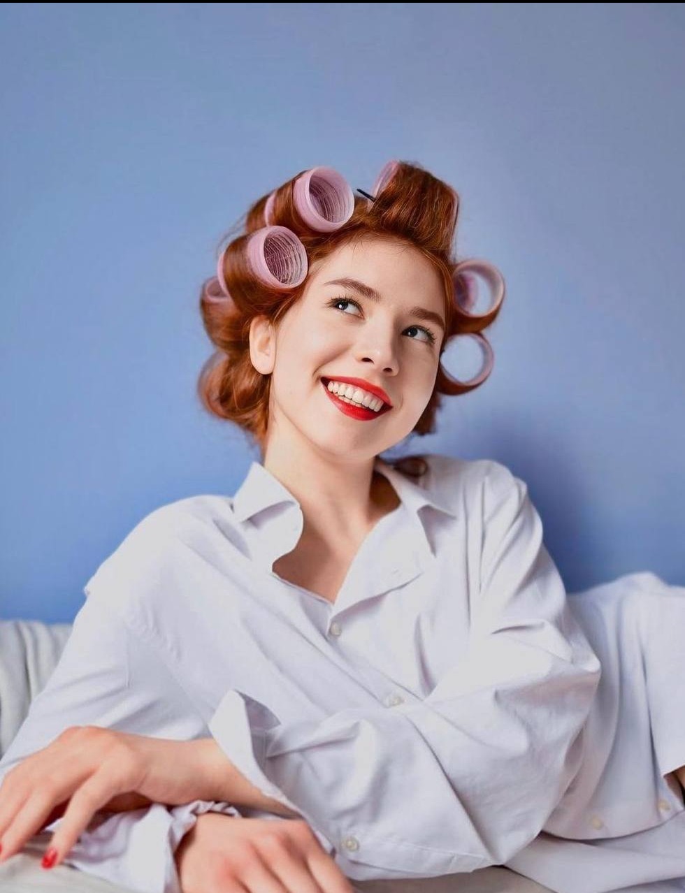 Uśmiechnięta, ruda kobieta z różowymi wałkami na włosach, ubrana w białą koszulę, spogląda w górę na tle błękitnej ściany.