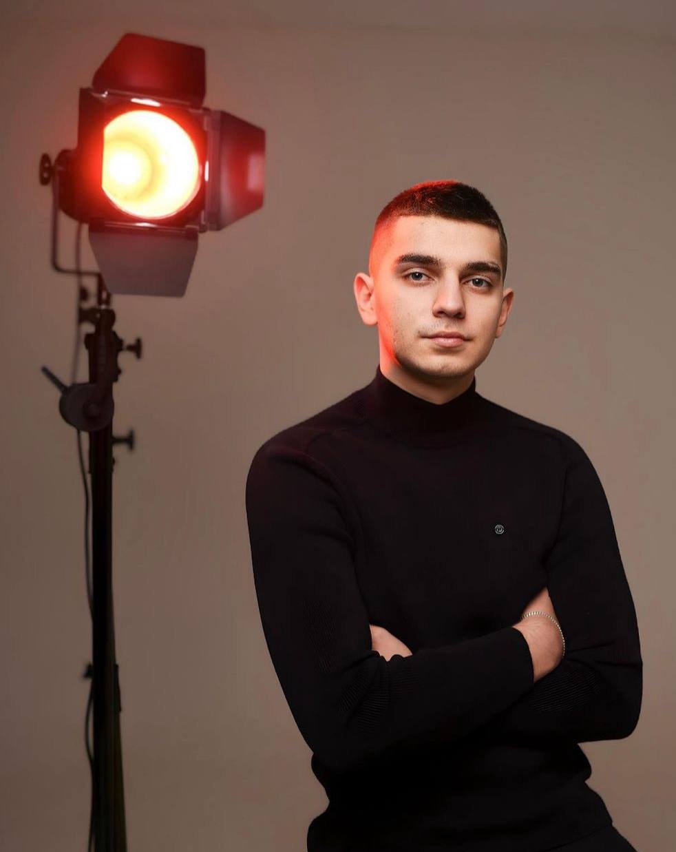 Portret młodego mężczyzny w czarnym golfie, oświetlonego czerwonym światłem studyjnym po lewej stronie, z rękami skrzyżowanymi na piersi.