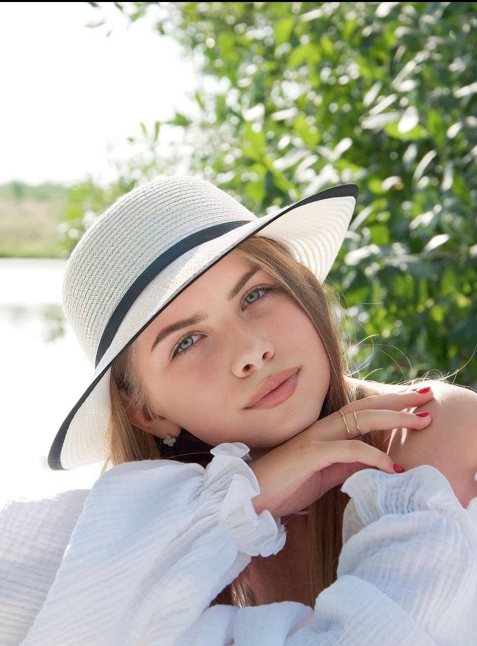 Portret młodej kobiety w białym kapeluszu z czarnym obramowaniem, opierającej głowę na dłoniach z czerwonym manicure na tle zieleni i wody.