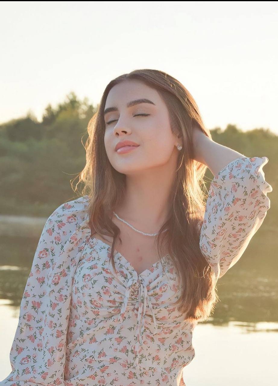 Portret młodej kobiety z zamkniętymi oczami, w bluzce w kwiaty i perłowym naszyjniku, z ręką uniesioną do włosów, na tle zieleni i wody w ciepłym świetle.