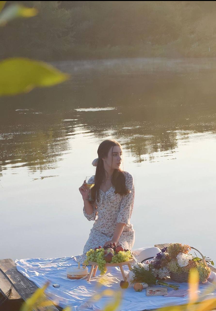 Młoda kobieta w kwiecistej sukience siedzi na pomoście nad jeziorem, trzyma kieliszek wina, obok rozłożony piknik z owocami i kwiatami, w tle lekka mgła nad wodą.