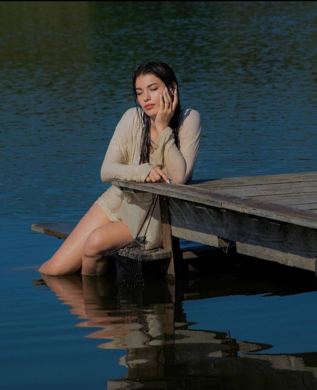 Młoda kobieta w jasnej sukience siedzi na drewnianym pomoście zanurzona w wodzie, zamyślona poza w letnim słońcu.