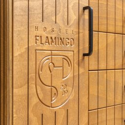 IXIDO Sp. z o.o. - Drewniana szafa z szufladami, z wygrawerowanym logo 'Hostel Flamingo' i rokiem 2009, z czarnymi uchwytami.