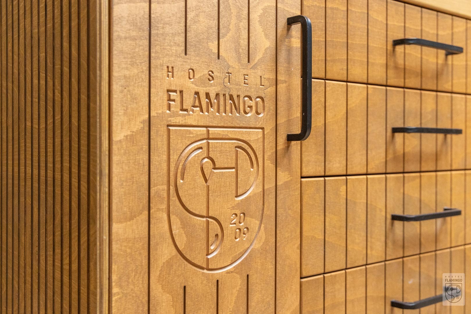 Drewniana szafa z szufladami, z wygrawerowanym logo 'Hostel Flamingo' i rokiem 2009, z czarnymi uchwytami.
