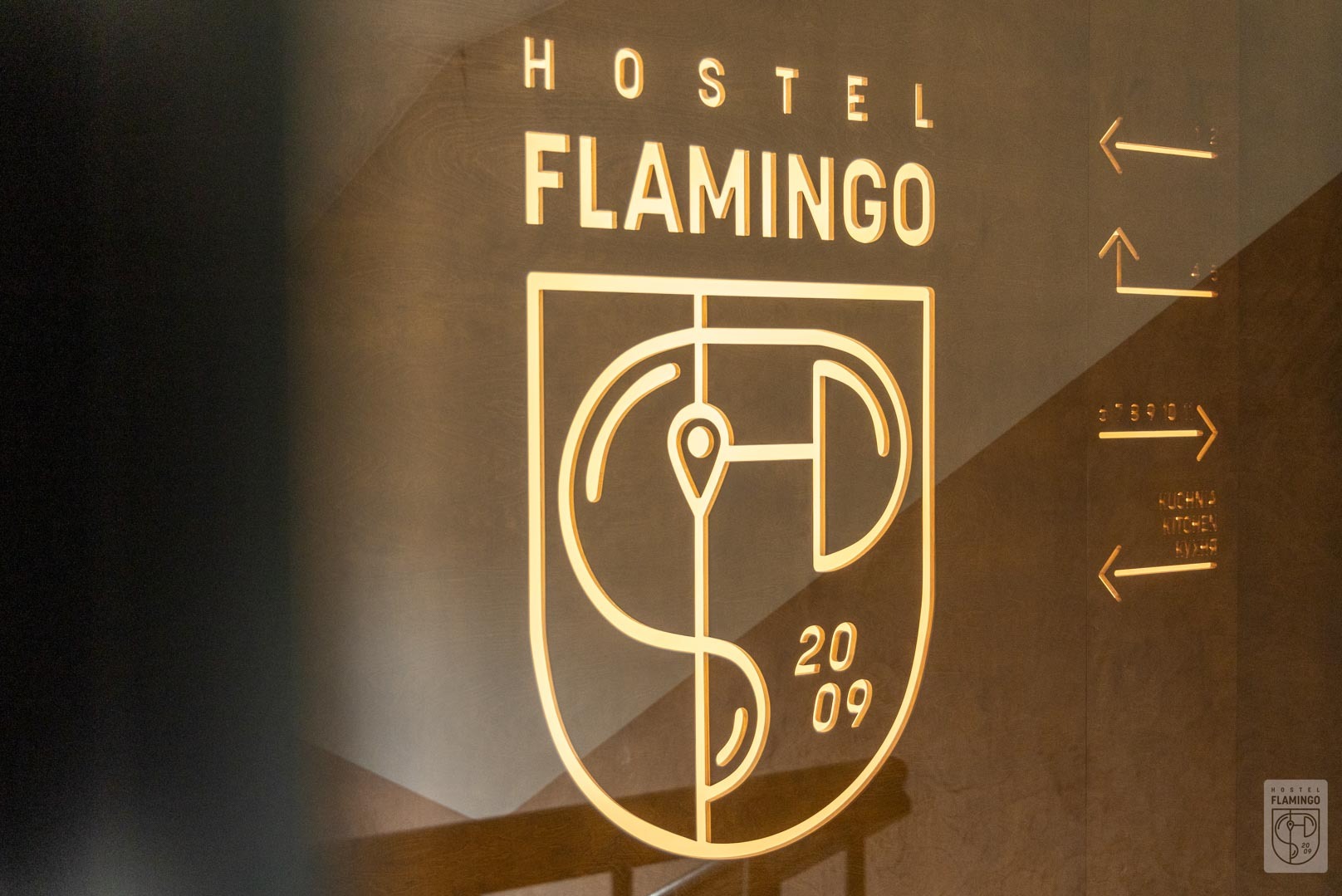 Podświetlany szyld Hostel Flamingo z logo, datą 2009 i wskazówkami kierunku do kuchni