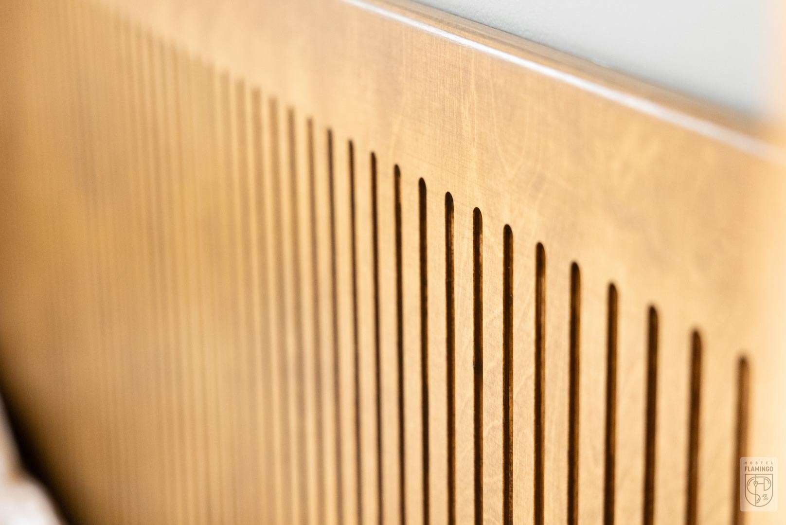 Drewniana płyta z pionowymi, równoległymi wycięciami, widoczna pod kątem, z logo hostelu w prawym dolnym rogu.