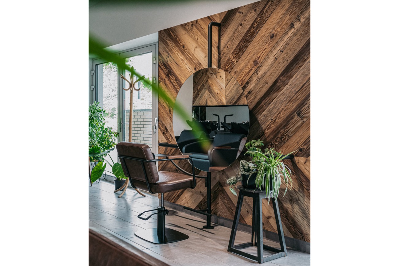 Nowoczesne wnętrze salonu fryzjerskiego z drewnianą ścianą w jodełkę, okrągłym lustrem i skórzanym fotelem fryzjerskim z metalową podstawą, uzupełnione roślinami doniczkowymi.