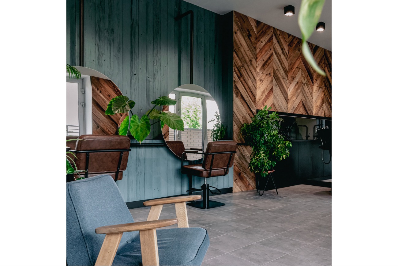 Wnętrze biura lub salonu z drewnianą ścianą w jodełkę, dwoma okrągłymi lustrami, skórzanymi fotelami fryzjerskimi i niebieskim fotelem wypoczynkowym.