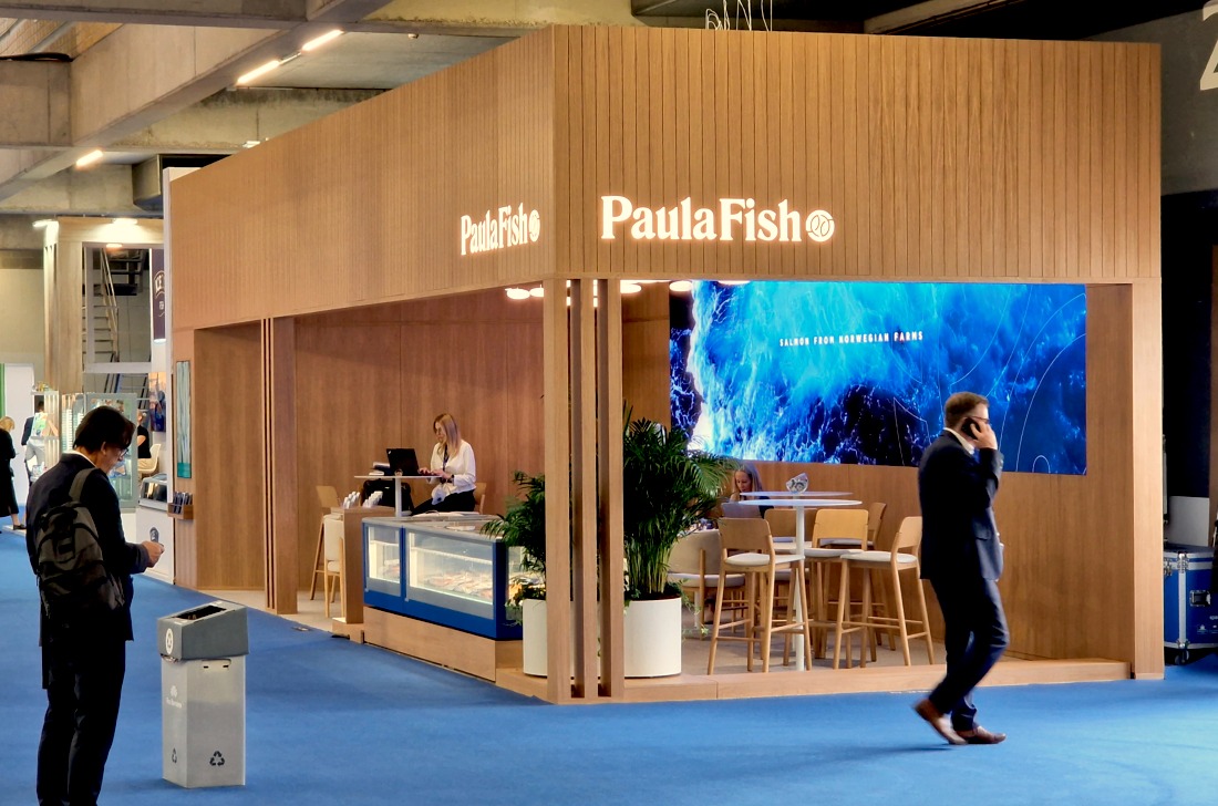 Stoisko targowe z drewnianą zabudową, logiem PaulaFish i ekranem LED wyświetlającym wizualizację oceanu. Osoby w garniturach i białych koszulach wewnątrz stoiska.