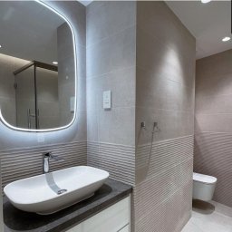 PRESKO MATEUSZ ŻMUDA - Nowoczesna łazienka z podświetlanym lustrem, umywalką nablatową i płytkami z falistym wzorem w odcieniach szarości, widoczna kabina prysznicowa w tle.