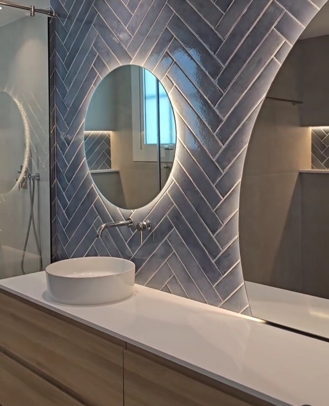 Nowoczesna łazienka z umywalką nablatową, podświetlanym lustrem owalnym i szarą glazurą ułożoną w jodełkę, z widoczną armaturą chromowaną.