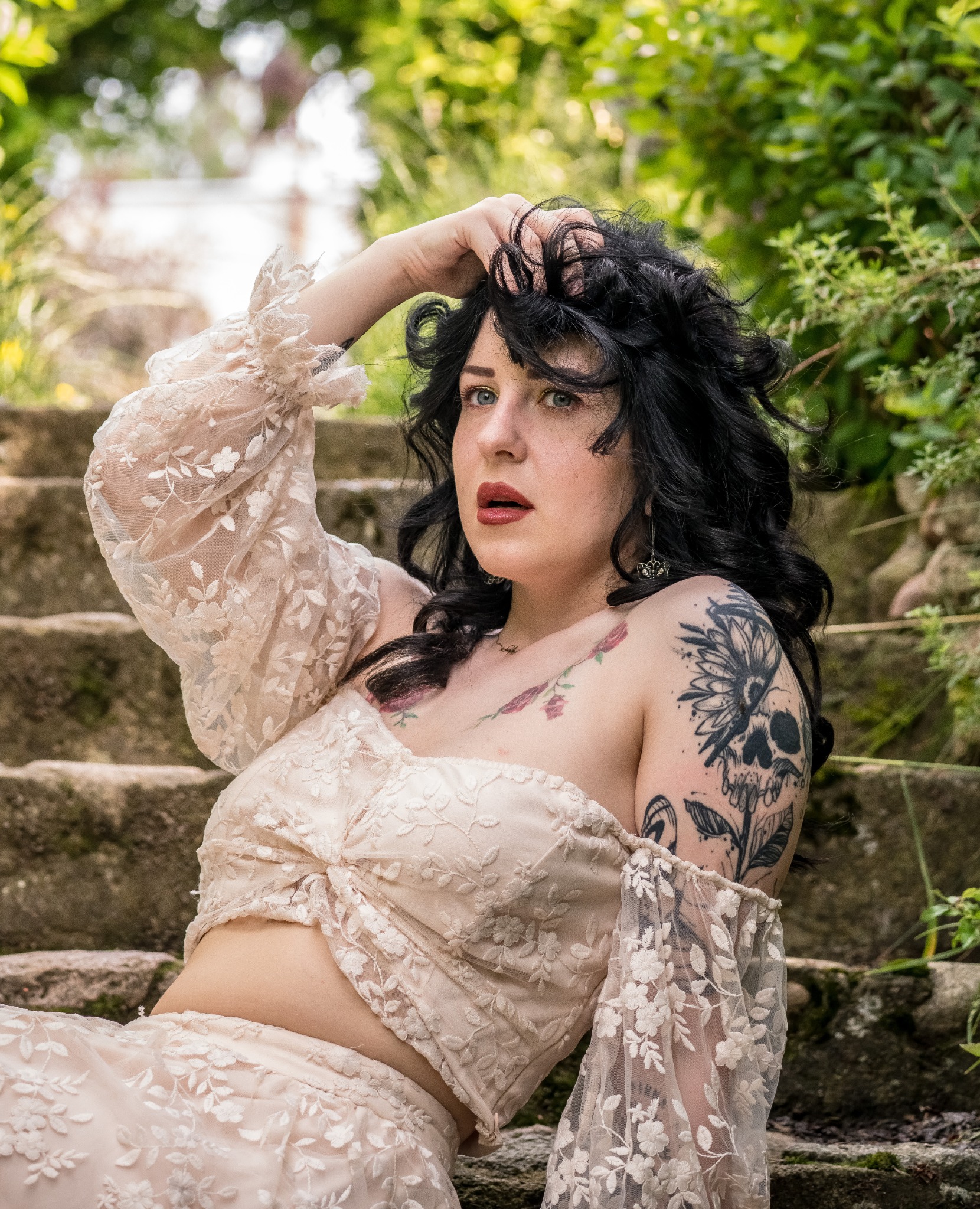 Kobieta w koronkowej sukni pozuje na kamiennych schodach, z tatuażami na ramionach i czarnymi włosami, trzymając rękę na głowie, otoczona zielenią.