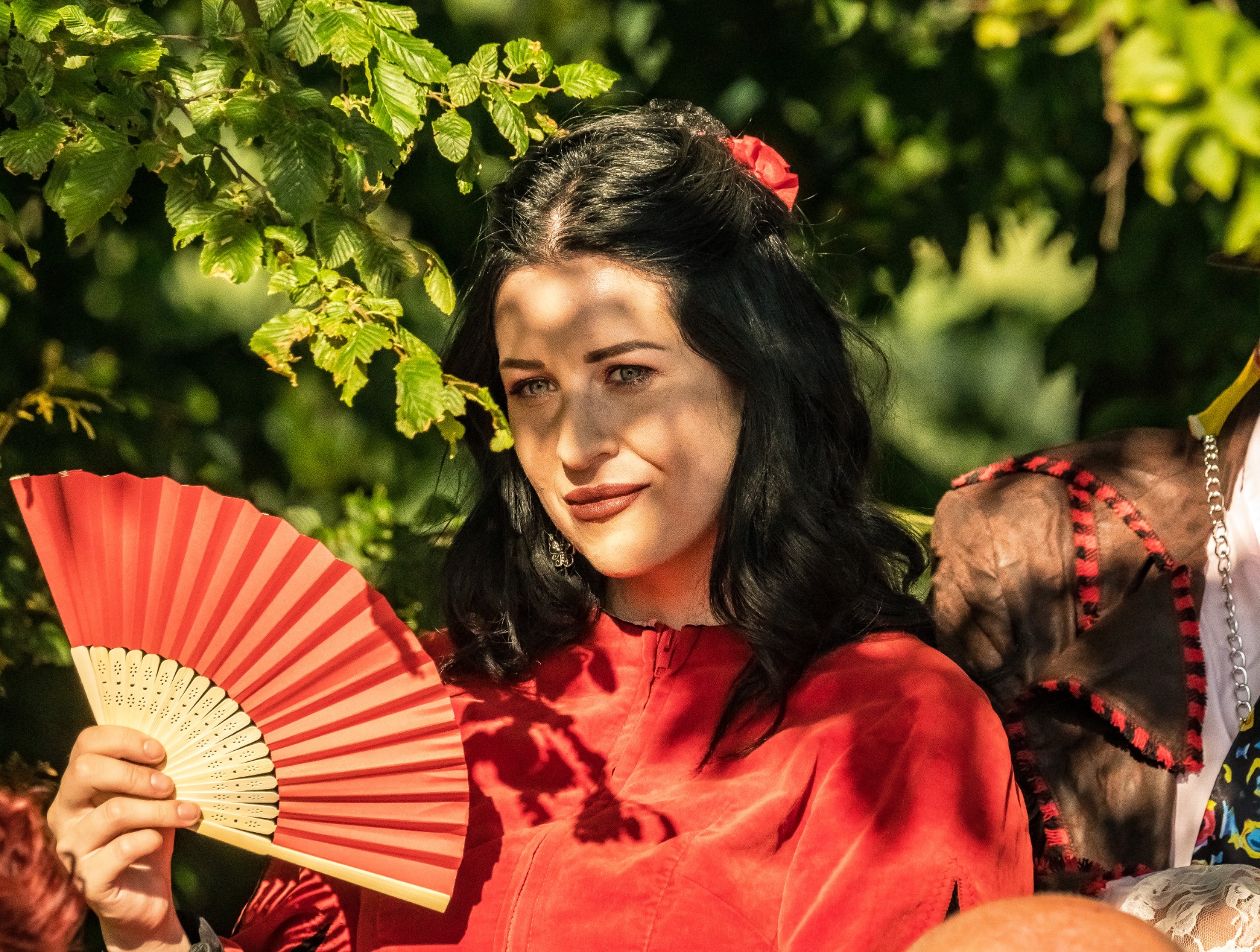 Kobieta w czerwonej sukience z wachlarzem na tle zieleni, cień na twarzy, czerwony kwiat we włosach, stylizacja retro.