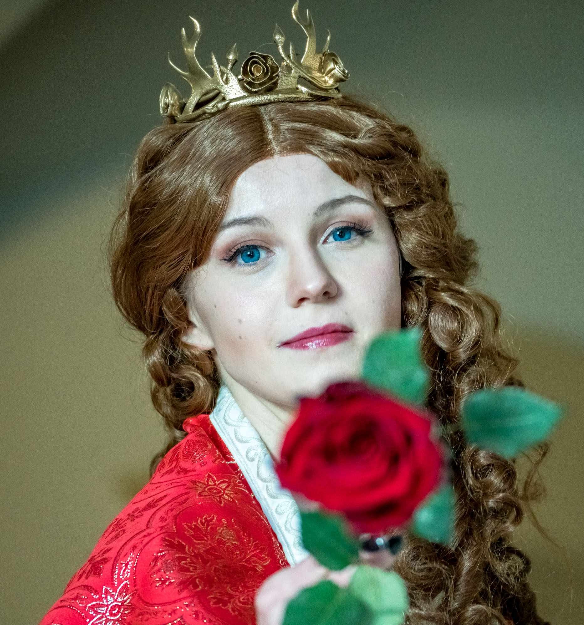 Portret kobiety z błękitnymi oczami, w złotej koronie i czerwonej sukni, trzymającej czerwoną różę na rozmytym tle.