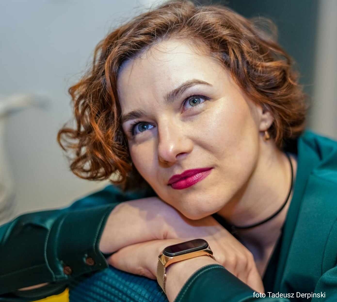 Portret kobiety o rudych kręconych włosach, w zielonej marynarce, z delikatnym makijażem i złotym zegarkiem, opierającej się na rękach i spoglądającej w górę.