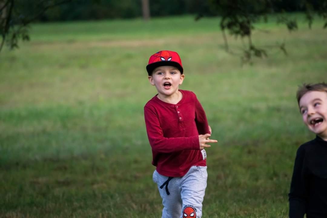 Chłopiec w czerwonej koszulce i czapce z motywem Spidermana biegnie po trawie, w tle drugi roześmiany chłopiec.