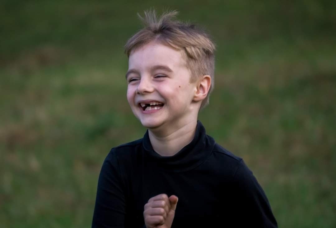 Uśmiechnięty chłopiec w czarnym golfie, z widocznymi brakami w uzębieniu, zaciśniętą dłonią, na rozmytym zielonym tle.