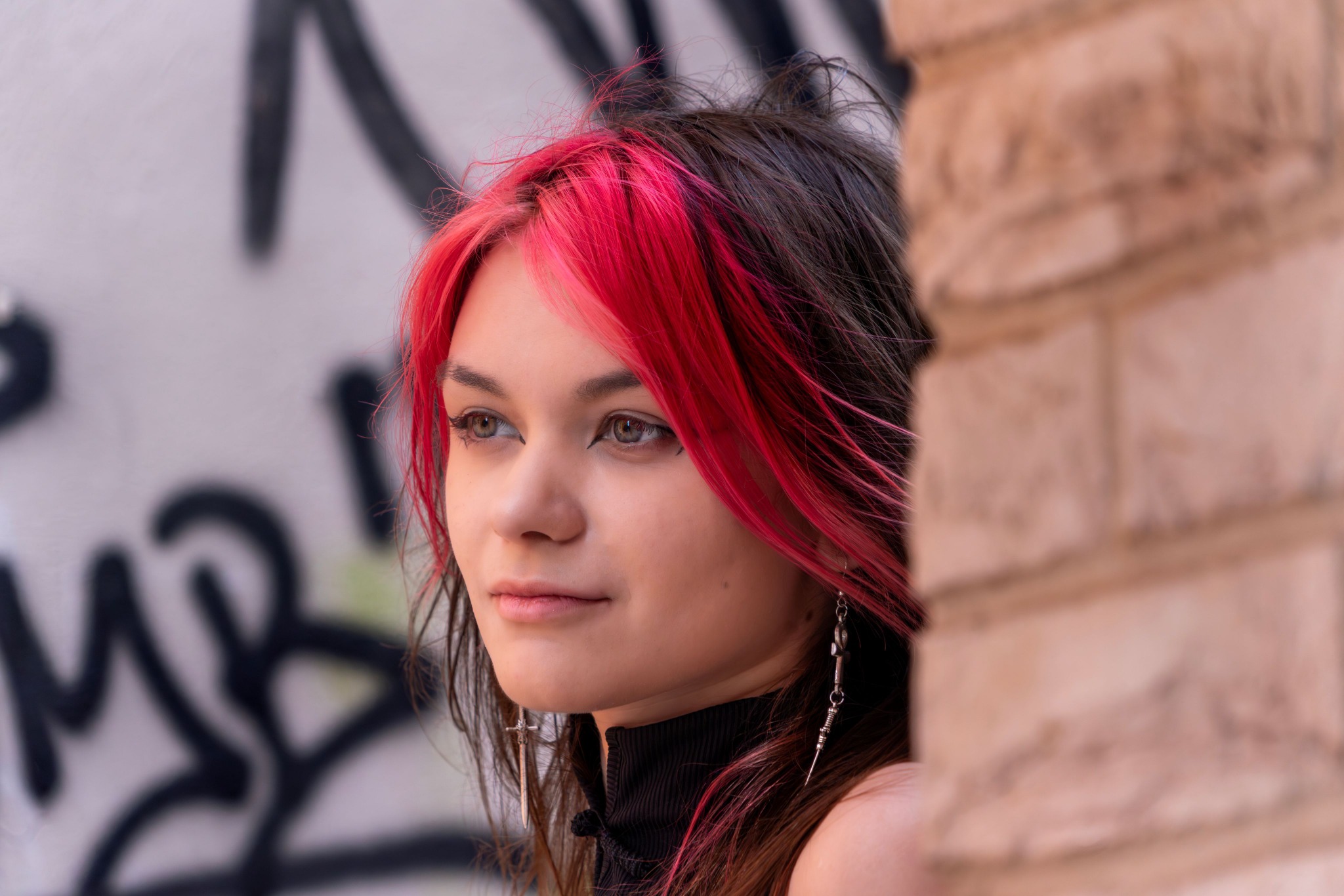 Portret młodej kobiety ze Szczecina z czerwonymi pasemkami i kolczykami na tle ściany z graffiti, patrzy w bok. Ujęcie z bliska, miękkie światło.