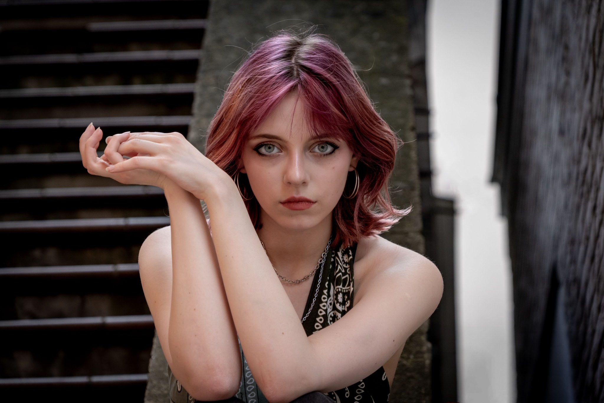 Portret młodej kobiety z różowymi włosami, siedzącej na tle betonowych schodów. Uwagę przykuwają duże kolczyki koła i wyraziste spojrzenie.