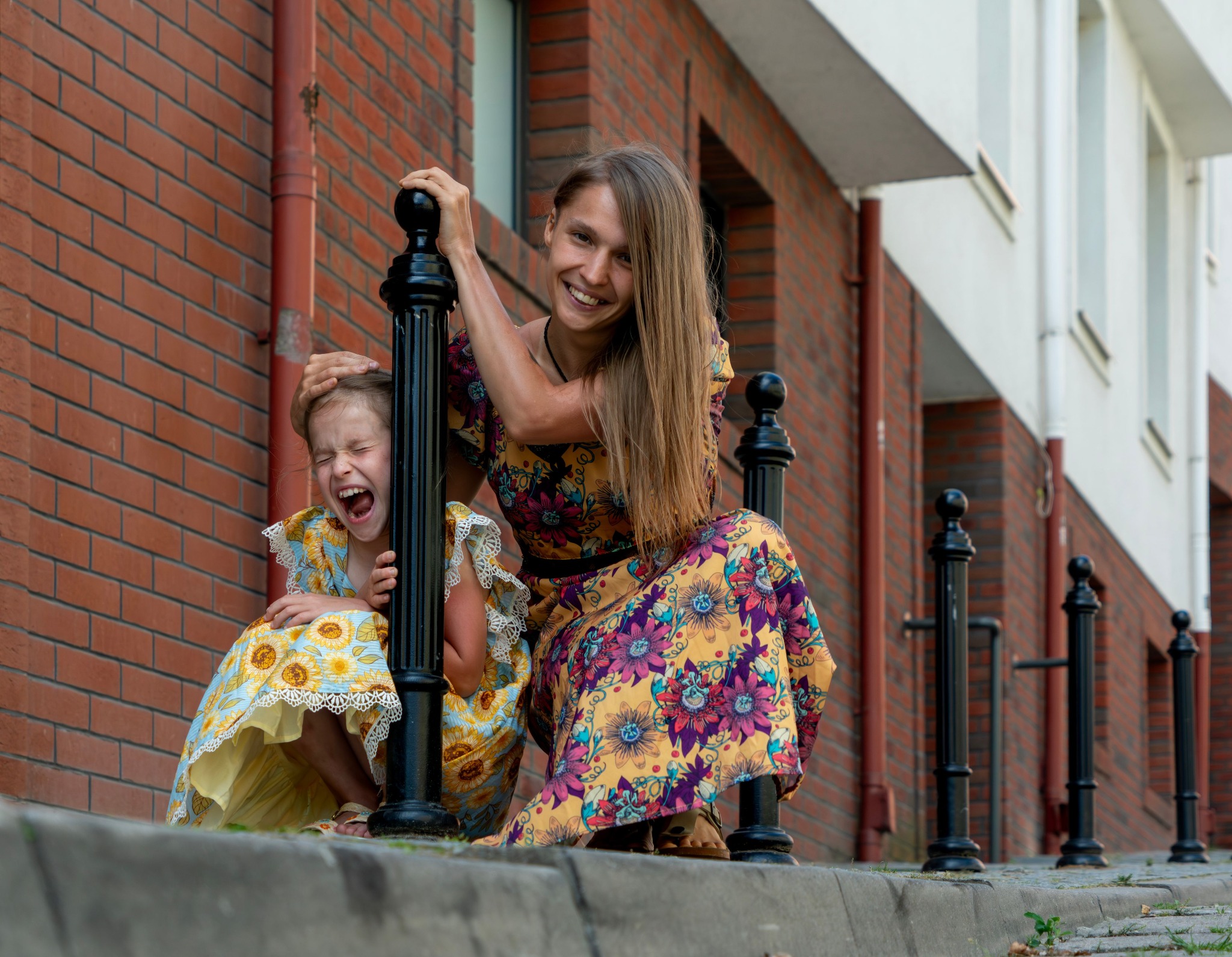 Uśmiechnięta mama i krzycząca córka w kwiecistych sukienkach pozują przy czarnych słupkach na tle ceglanej ściany budynku.