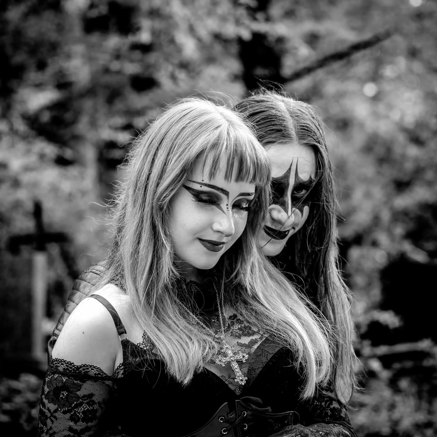 Czarno-biała sesja gotycka: kobieta z blond włosami i mężczyzna z makijażem klauna na tle drzew i krzyża na cmentarzu. Mroczny klimat, detale stroju i makijażu.