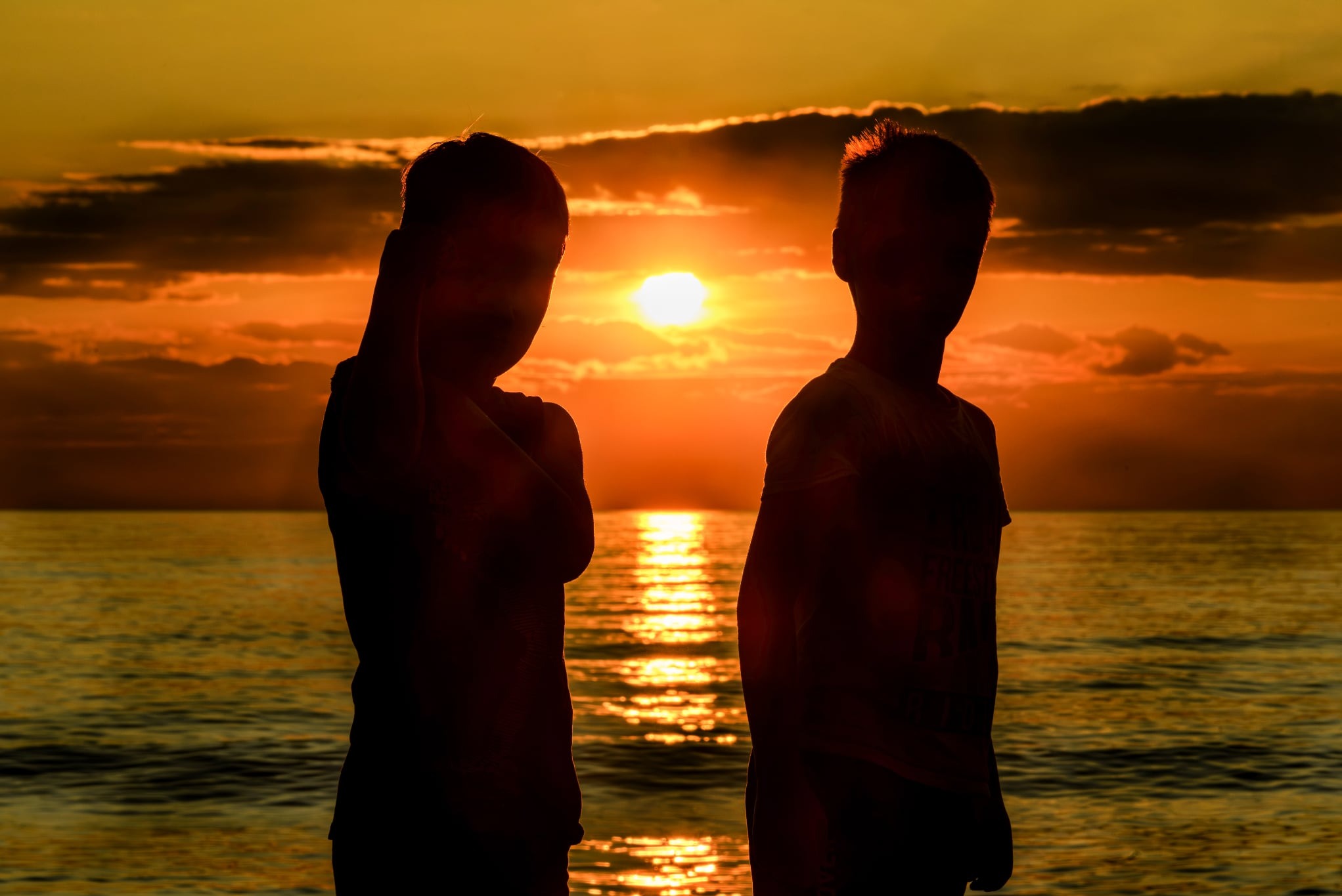 Sylwetki dwóch osób na tle zachodzącego słońca nad morzem, tworzące ciepły, kontrastowy obraz. Słońce odbija się w wodzie, dodając blasku scenie.