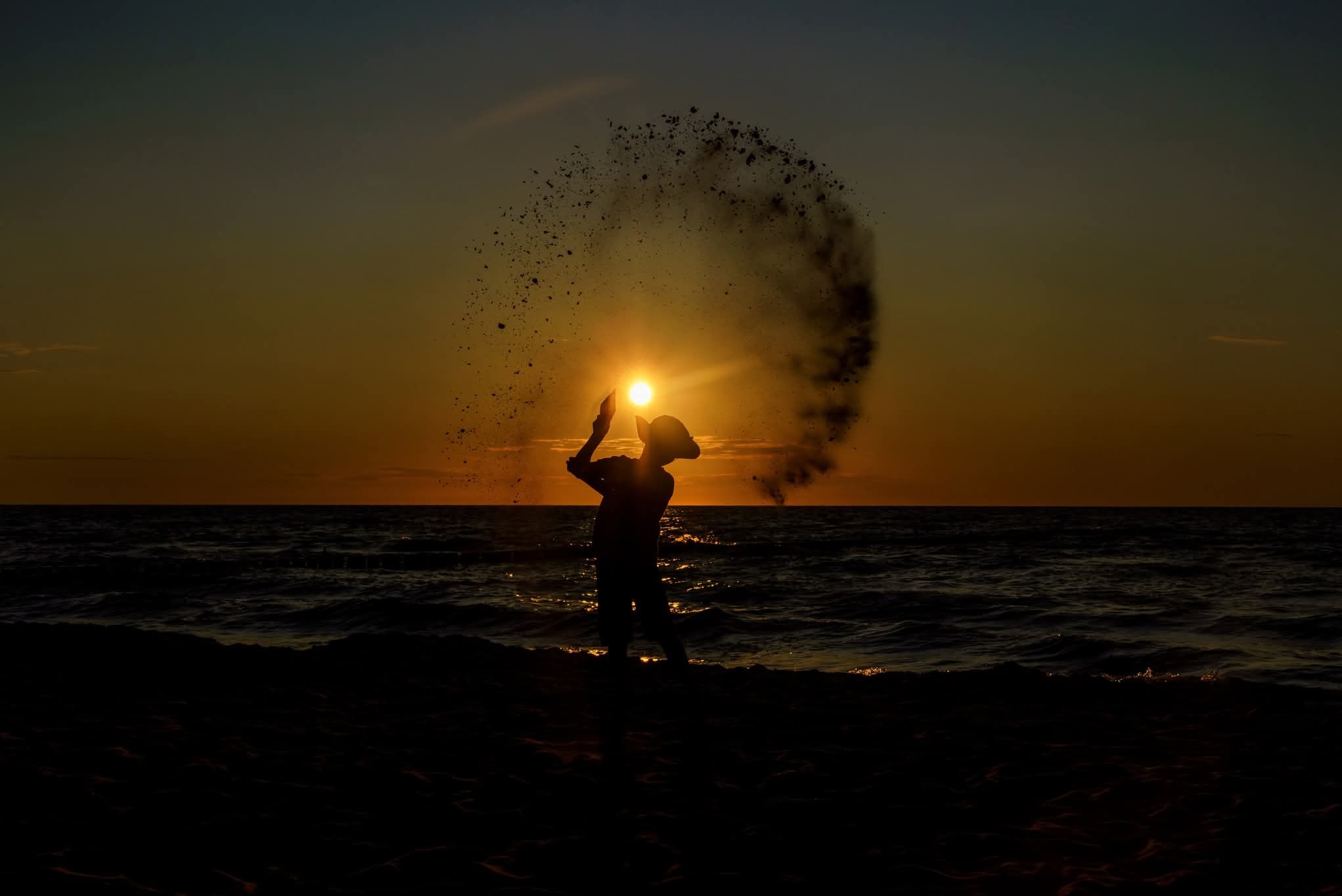 Sylwetka osoby rzucającej piasek w górę na tle zachodzącego słońca nad morzem. Ciepłe barwy, dynamiczne ujęcie, plaża.
